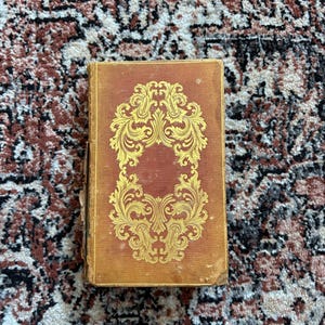 May include: A vintage book with a worn, reddish-brown cover. The cover features an ornate gold design in the center. The book is rectangular and appears to be in good condition, with slight wear on the edges. The background is a patterned rug.