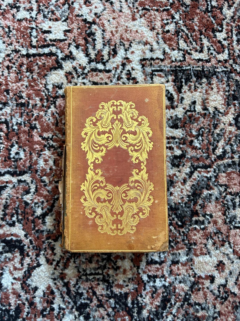 May include: A vintage book with a worn, reddish-brown cover. The cover features an ornate gold design in the center. The book is rectangular and appears to be in good condition, with slight wear on the edges. The background is a patterned rug.