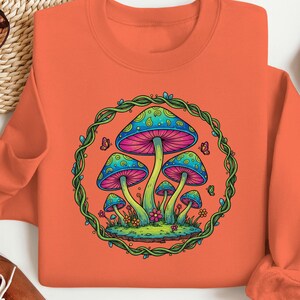 May include: Orange crewneck sweatshirt featuring a colorful mushroom graphic. The design includes blue, pink, and green mushrooms within a green vine border, with small butterflies and flowers. The sweatshirt is made of a soft material.