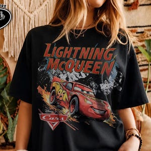 Peut inclure: Un t-shirt noir &agrave; manches courtes avec une impression graphique de la voiture de course rouge Flash McQueen du film Cars. Le texte Lightning McQueen est imprim&eacute; en grandes lettres rouges au-dessus de la voiture, avec le logo Cars en dessous.