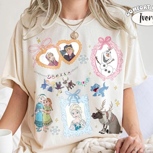 May include: An ivory short-sleeve t-shirt featuring a graphic print of characters from the movie Frozen. The design includes framed portraits of Anna, Kristoff, Elsa, and Olaf, along with illustrations of Sven and Bruni, plus the text Frozen.