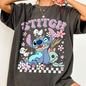 May include: A charcoal grey t-shirt featuring a graphic print of the character Stitch with a small duck on its head, another duck to the side, and a doll. The text STITCH is printed above in purple, surrounded by pink and yellow flowers and a checkerboard pattern.