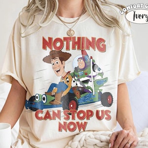 K&ouml;nnte beinhalten: Ein elfenbeinfarbenes Kurzarm-T-Shirt mit einer Grafik von Woody und Buzz Lightyear in einem blauen Rennwagen. Der Text dar&uuml;ber lautet NOTHING und darunter CAN STOP US NOW. Ein kleines rundes Etikett in der Ecke zeigt COMFORT COLOR IVORY.