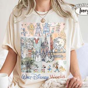 May include: An ivory t-shirt featuring a colorful graphic print of Walt Disney World landmarks and characters including Mickey Mouse, Minnie Mouse, Donald Duck, Goofy, and Pluto. The text reads Walt Disney World and Comfort Color Ivory.