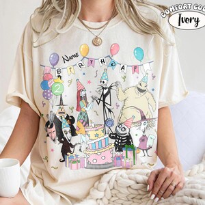 May include: An ivory t-shirt featuring a graphic print of characters from The Nightmare Before Christmas at a birthday party. The design includes balloons, a tiered cake, presents, and a banner with the text Name BIRTHDAY. A Comfort Color Ivory label is visible.
