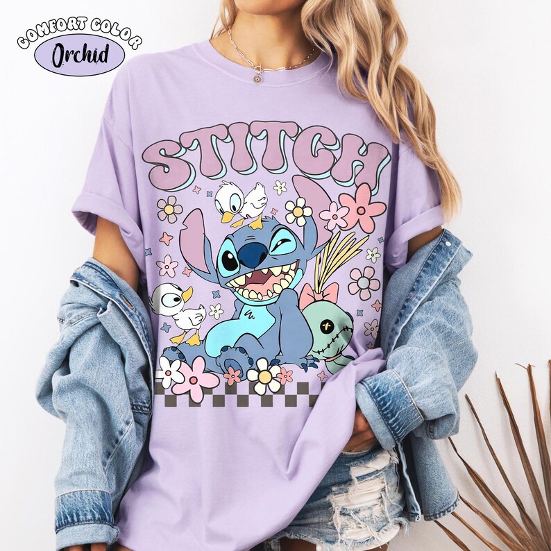 May include: An orchid-coloured t-shirt featuring a graphic of the character Stitch, two small white ducks, and a doll. The text STITCH is printed in large purple letters above the character. A small label in the corner reads COMFORT COLOR Orchid.