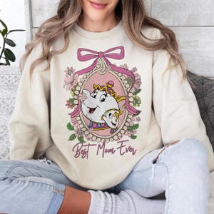 May include: Cream-colored sweatshirt featuring a cartoon image of Mrs. Potts and Chip from Beauty and the Beast, framed by a pink oval and floral accents. The text "Best Mom Ever" is written below the image.