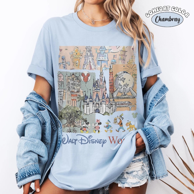 May include: A light blue t-shirt featuring a graphic print of Walt Disney World landmarks and characters. The design includes a castle, a sphere, and various cartoon figures. The text Walt Disney World is printed at the bottom of the graphic.