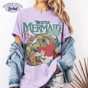 Op de afbeelding: Een orchideekleurig t-shirt met een grafische print van De Kleine Zeemeermin met Botje en Sebastiaan bij een onderwaterkasteel. De tekst luidt The Little Mermaid. Een blauw spijkerjack wordt over het t-shirt gedragen.