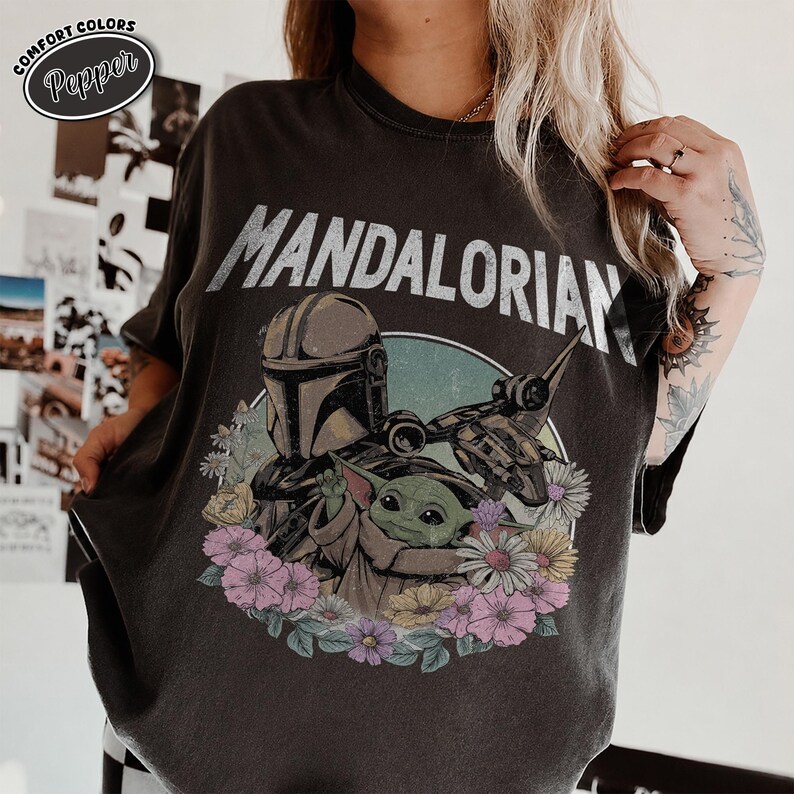 K&ouml;nnte beinhalten: Ein anthrazitfarbenes Kurzarm-T-Shirt mit einem Grafikdruck des Mandalorianers und Grogu, umgeben von bunten Blumen. Der Text "MANDALORIAN" ist in gro&szlig;en wei&szlig;en Buchstaben &uuml;ber den Figuren aufgedruckt. Ein "COMFORT COLORS Pepper"-Etikett ist sichtbar.