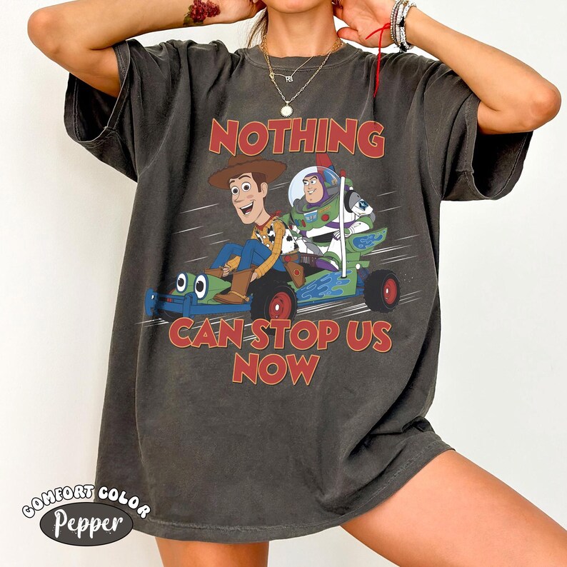 K&ouml;nnte beinhalten: Ein anthrazitfarbenes Kurzarm-T-Shirt mit einer Grafik von Woody und Buzz Lightyear auf einem gr&uuml;nen und blauen Go-Kart. Der Text "NOTHING CAN STOP US NOW" ist in roter, fetter serifenloser Schrift &uuml;ber und unter den Figuren aufgedruckt.