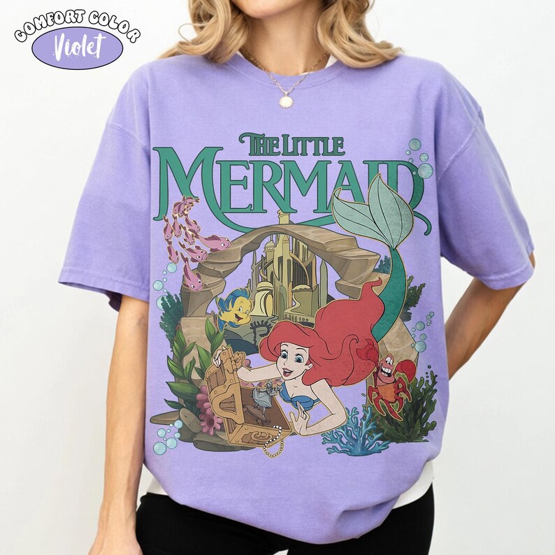 Op de afbeelding: Een violetkleurig T-shirt met korte mouwen en een grafische print van De Kleine Zeemeermin. Het ontwerp toont Ari&euml;l, Botje en Sebastiaan bij een schatkist en een onderwaterkasteel. De tekst luidt The Little Mermaid in groene letters.
