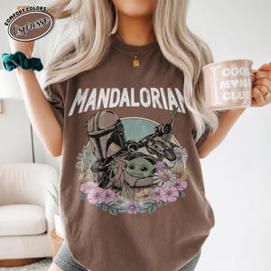 K&ouml;nnte beinhalten: Ein espressofarbenes T-Shirt mit dem Schriftzug MANDALORIAN in wei&szlig;en Buchstaben &uuml;ber einer Grafik des Mandalorianers und Grogu, umgeben von bunten Blumen. In einer Hand wird eine hellrosa Tasse mit dem Text COOL MOM CLUB gehalten.
