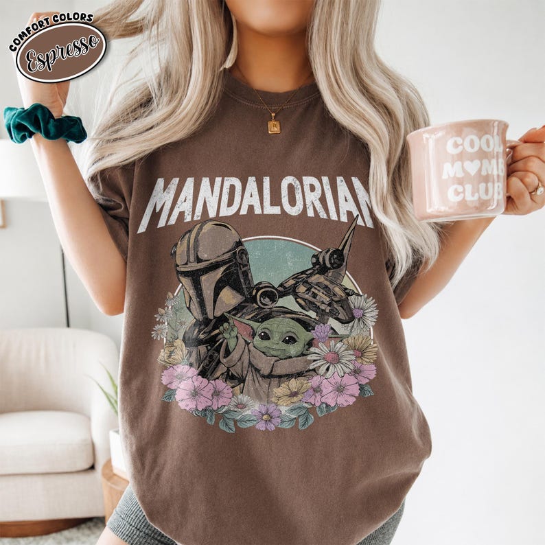 K&ouml;nnte beinhalten: Ein espressofarbenes T-Shirt mit dem Schriftzug MANDALORIAN in wei&szlig;en Buchstaben &uuml;ber einer Grafik des Mandalorianers und Grogu, umgeben von bunten Blumen. In einer Hand wird eine hellrosa Tasse mit dem Text COOL MOM CLUB gehalten.