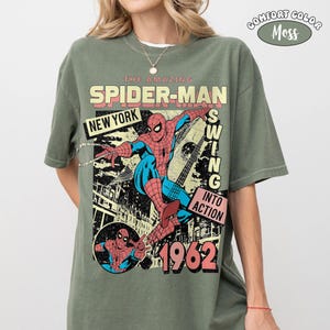 May include: A moss green short-sleeve t-shirt featuring a vintage-style graphic of Spider-Man in red and blue. The print includes the text THE AMAZING SPIDER-MAN, NEW YORK, SWING INTO ACTION, and 1962. A small circular logo reads COMFORT COLOR MOSS.