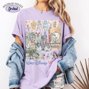 May include: An orchid-colored t-shirt featuring a graphic print of Walt Disney World landmarks and characters. The design includes sketches of the castle, Epcot sphere, and various cartoon figures, with the text Walt Disney World printed below.