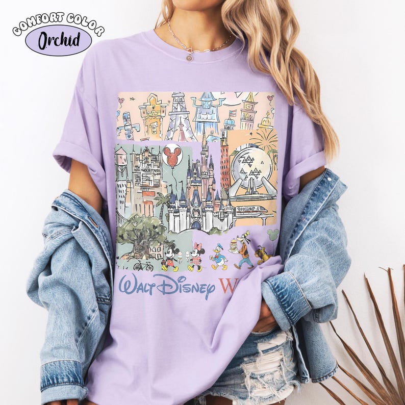 May include: An orchid-colored t-shirt featuring a graphic print of Walt Disney World landmarks and characters. The design includes sketches of the castle, Epcot sphere, and various cartoon figures, with the text Walt Disney World printed below.