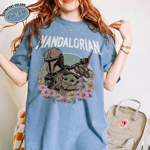 K&ouml;nnte beinhalten: Ein jeansblaues T-Shirt mit einer Grafik des Mandalorianers und Grogu, umgeben von bunten Blumen. Der Text "MANDALORIAN" ist in einer abgenutzten wei&szlig;en Schrift &uuml;ber der Illustration gedruckt. Ein "Comfort Colors"-Logo befindet sich in der Ecke.