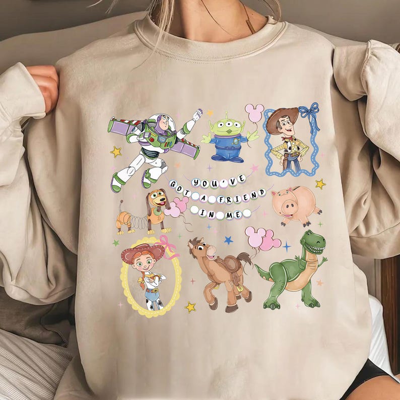 May include: A beige sweatshirt featuring colorful cartoon characters from the Toy Story movie. The design includes Buzz Lightyear, Woody, Jessie, an alien, a dinosaur, and the phrase "You've Got a Friend in Me."
