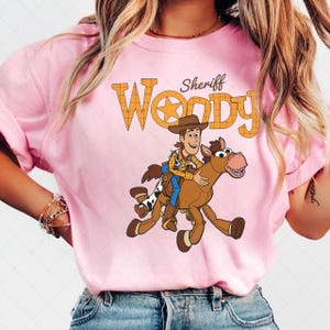 May include: Pink t-shirt featuring a cartoon illustration of Sheriff Woody riding a horse. The text "Woody" is displayed in orange with a star in the center. The word "Sheriff" is above the word "Woody".