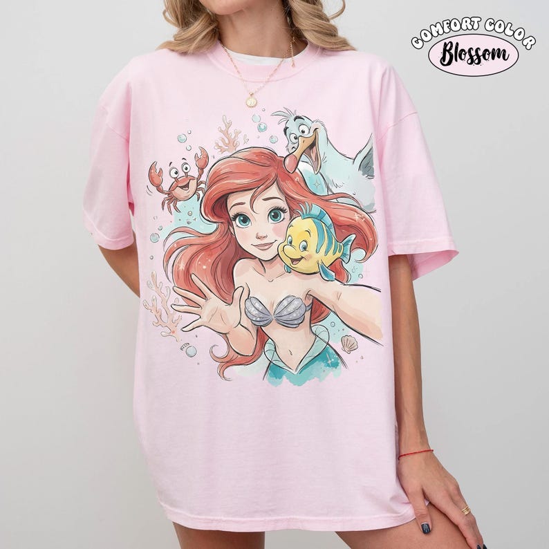 May include: A light pink t-shirt featuring a graphic print of a red-haired mermaid, a yellow and blue fish, a small red crab, and a white seagull. The design includes bubbles and coral elements. The text "COMFORT COLOR Blossom" appears in the top right corner.