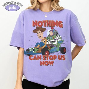 K&ouml;nnte beinhalten: Ein violettes Kurzarm-T-Shirt mit einer Grafik von Woody und Buzz Lightyear auf einem gr&uuml;nen Go-Kart. Der Text &uuml;ber und unter den Figuren lautet "NOTHING CAN STOP US NOW". Ein kleines Logo oben links zeigt "COMFORT COLOR Violet".