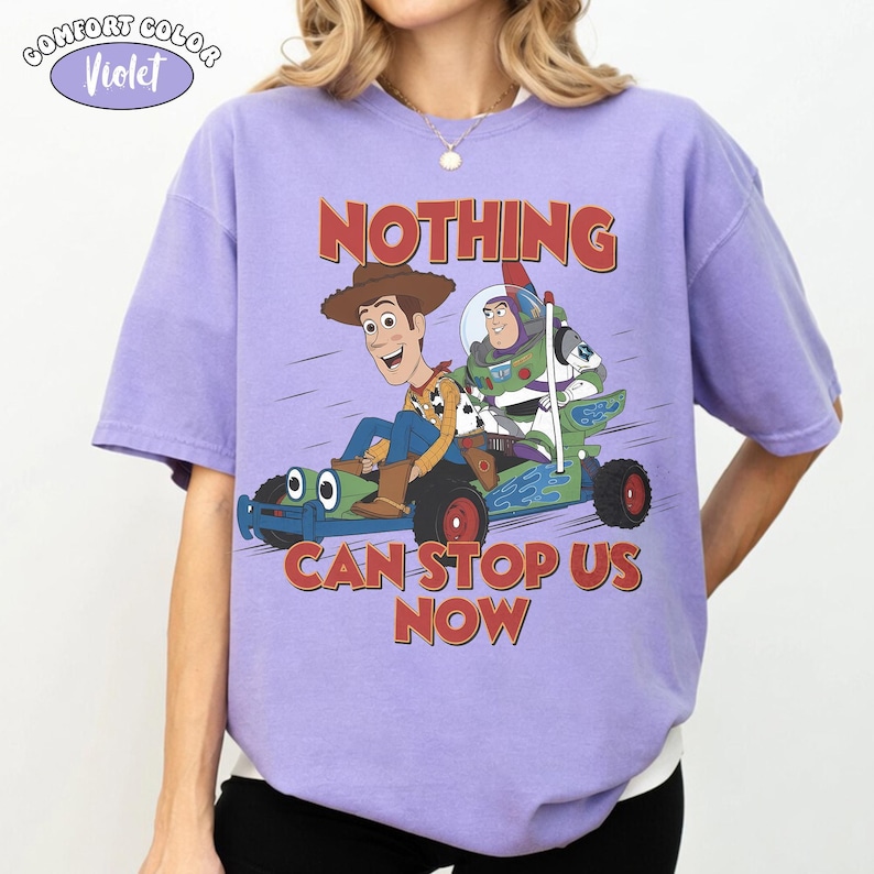 K&ouml;nnte beinhalten: Ein violettes Kurzarm-T-Shirt mit einer Grafik von Woody und Buzz Lightyear auf einem gr&uuml;nen Go-Kart. Der Text &uuml;ber und unter den Figuren lautet "NOTHING CAN STOP US NOW". Ein kleines Logo oben links zeigt "COMFORT COLOR Violet".