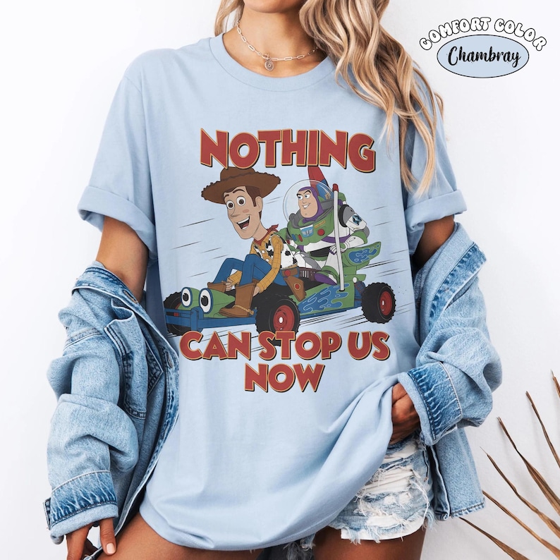K&ouml;nnte beinhalten: Ein hellblaues T-Shirt mit kurzen &Auml;rmeln und einer Grafik von Woody und Buzz Lightyear in einem Go-Kart. Der Text lautet NOTHING CAN STOP US NOW. Ein kreisf&ouml;rmiges Logo oben rechts zeigt den Text COMFORT COLOR Chambray.
