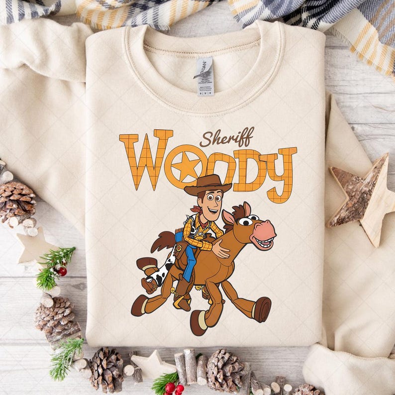May include: Cream-colored sweatshirt featuring a graphic of Sheriff Woody riding a horse. The text "Sheriff Woody" is printed above the illustration in orange and brown. The sweatshirt has a crew neck and long sleeves.