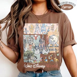 May include: An espresso brown t-shirt featuring a graphic print of various theme park landmarks, including a castle, a geodesic sphere, and a tower. The design includes cartoon characters walking at the bottom and the text Walt Disney World.