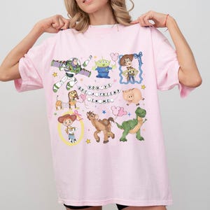 May include: A light pink t-shirt featuring colorful cartoon characters and the text "YOU'VE GOT A FRIEND IN ME." The design includes Buzz Lightyear, an alien, Woody, a pig, a dinosaur, and other characters.