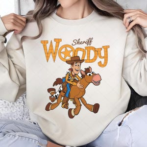 May include: Cream-colored sweatshirt featuring a cartoon illustration of Sheriff Woody riding a horse. The text "Sheriff Woody" is printed above the image in orange, plaid-patterned letters. The design is centered on the front of the sweatshirt.