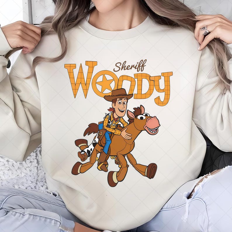 May include: Cream-colored sweatshirt featuring a cartoon illustration of Sheriff Woody riding a horse. The text "Sheriff Woody" is printed above the image in orange, plaid-patterned letters. The design is centered on the front of the sweatshirt.