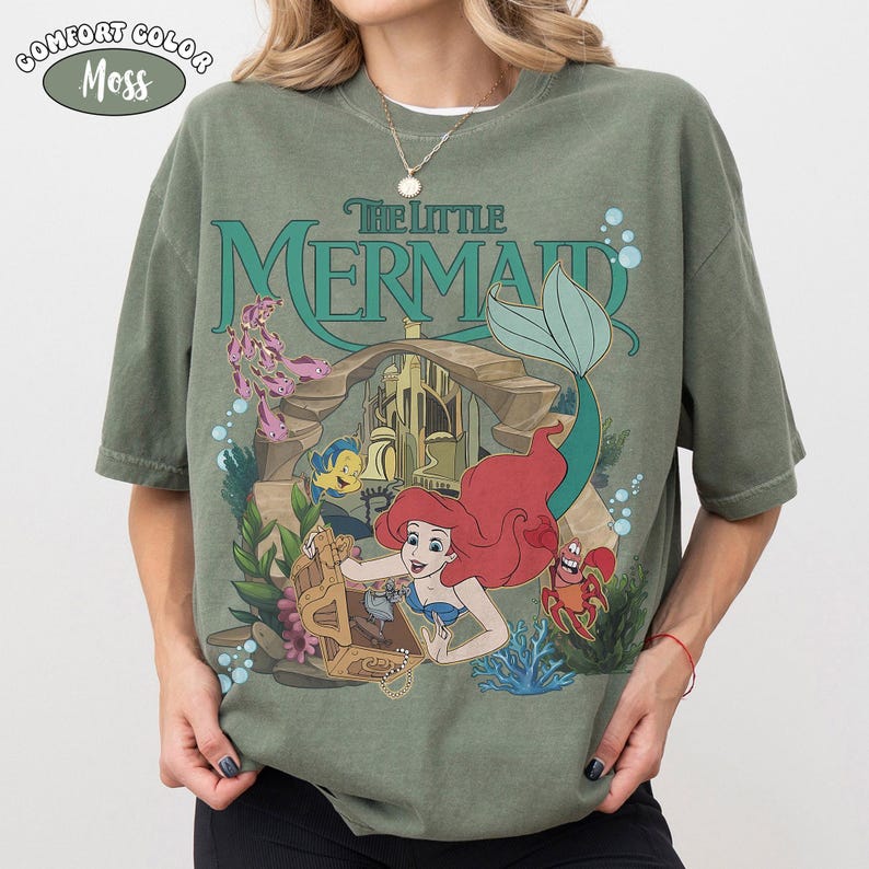 Op de afbeelding: Een mosgroen T-shirt met korte mouwen en een grafische print van De Kleine Zeemeermin. Het ontwerp toont Ari&euml;l, Botje en Sebastiaan bij een schatkist en een onderwaterkasteel. De tekst luidt The Little Mermaid in een blauwgroene kleur.