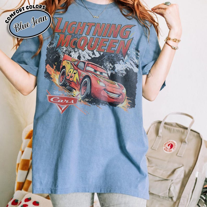 Peut inclure: Un t-shirt bleu jean avec une impression graphique de la voiture de course rouge Flash McQueen du film Cars. Le texte Lightning McQueen est imprim&eacute; en grandes lettres rouges au-dessus de la voiture, avec le logo Cars et le num&eacute;ro 95.