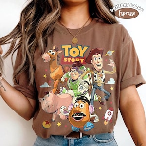 Puede incluir: Una camiseta color marr&oacute;n espresso con un estampado gr&aacute;fico de los personajes de Toy Story, incluyendo a Woody, Buzz Lightyear, Perdig&oacute;n, Hamm y el Se&ntilde;or Patata. El dise&ntilde;o incluye el texto Toy Story, un cohete peque&ntilde;o, un planeta y estrellas amarillas.
