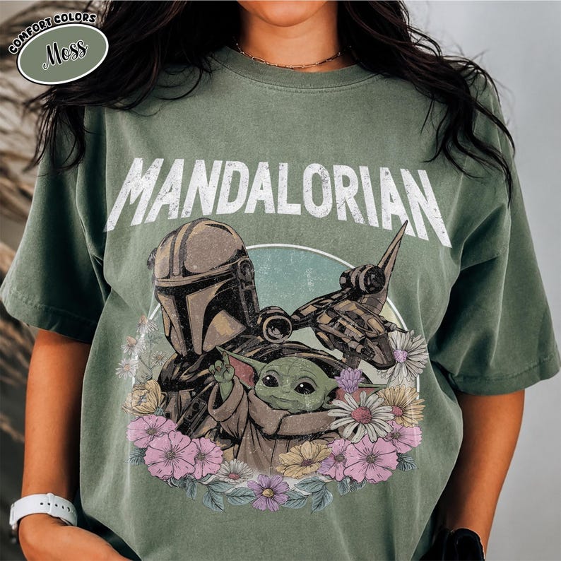 K&ouml;nnte beinhalten: Ein moosgr&uuml;nes T-Shirt mit einem Grafikdruck des Mandalorianers und Grogu, umgeben von bunten Blumen. Der Text "MANDALORIAN" ist in gro&szlig;en wei&szlig;en Buchstaben &uuml;ber den Figuren gedruckt. Das T-Shirt hat einen Rundhalsausschnitt und kurze &Auml;rmel.
