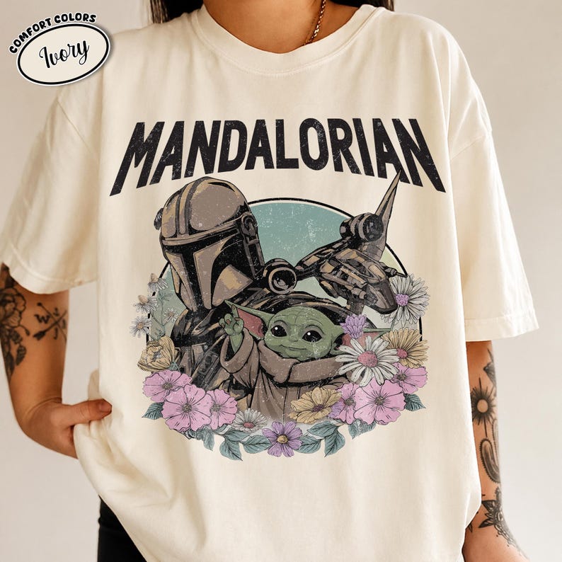 K&ouml;nnte beinhalten: Ein elfenbeinfarbenes Kurzarm-T-Shirt mit dem Text MANDALORIAN in schwarzen Buchstaben. Die Grafik zeigt eine Figur mit Helm und ein kleines gr&uuml;nes Wesen, umgeben von einer kreisf&ouml;rmigen Anordnung bunter Blumen und einer Raumschiff-Silhouette.