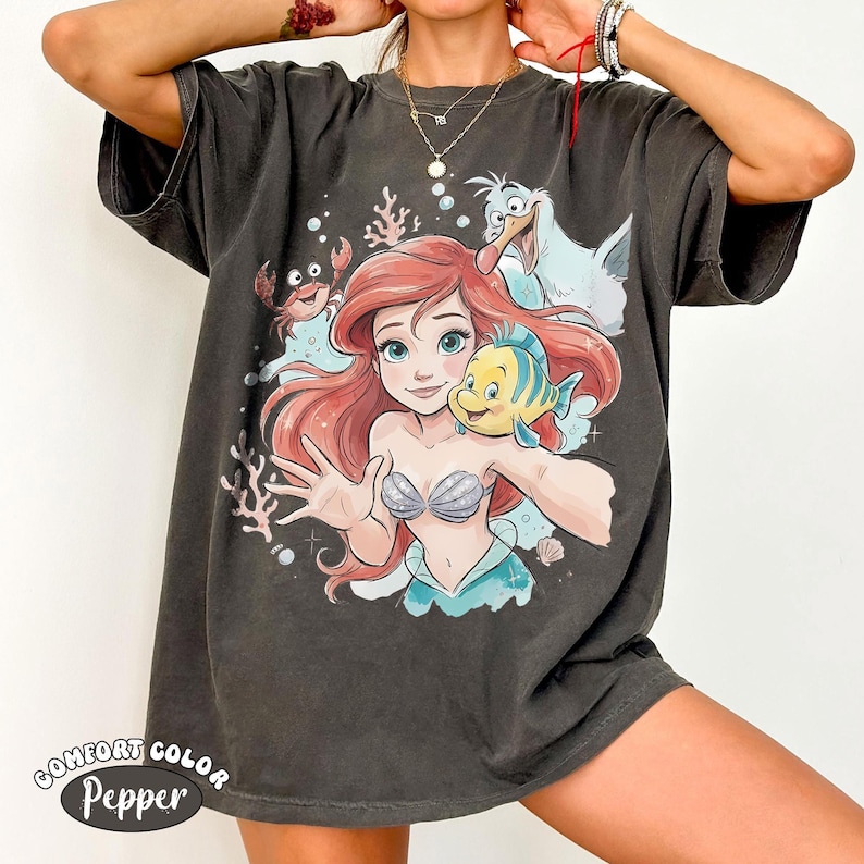 May include: A charcoal gray short-sleeve t-shirt featuring a graphic print of a red-haired mermaid character, a yellow and blue fish, a crab, and a white seagull. The design includes coral and bubble accents. A logo reads Comfort Color Pepper.