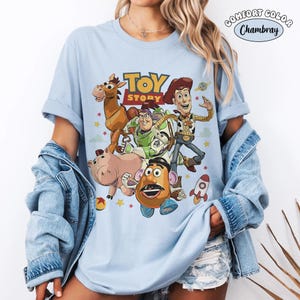 Puede incluir: Una camiseta azul claro con un estampado gr&aacute;fico de los personajes de Toy Story, incluyendo a Woody, Buzz Lightyear, Perdig&oacute;n, Hamm y el Sr. Patata. El dise&ntilde;o incluye el logotipo de Toy Story, un cohete y estrellas. Se ve una etiqueta Comfort Color Chambray.