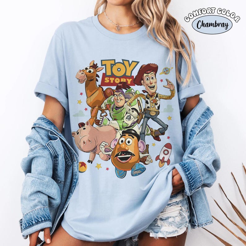 Puede incluir: Una camiseta azul claro con un estampado gr&aacute;fico de los personajes de Toy Story, incluyendo a Woody, Buzz Lightyear, Perdig&oacute;n, Hamm y el Sr. Patata. El dise&ntilde;o incluye el logotipo de Toy Story, un cohete y estrellas. Se ve una etiqueta Comfort Color Chambray.