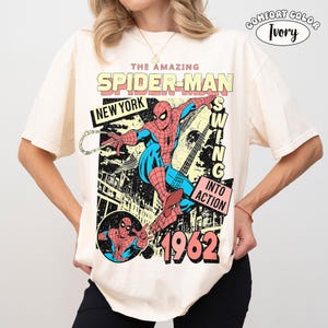 May include: An ivory t-shirt featuring a graphic print of Spider-Man in red and blue. The text reads THE AMAZING SPIDER-MAN, NEW YORK, SWING INTO ACTION, and 1962. A smaller circular graphic shows Spider-Man in a pose. The shirt has a crew neck.