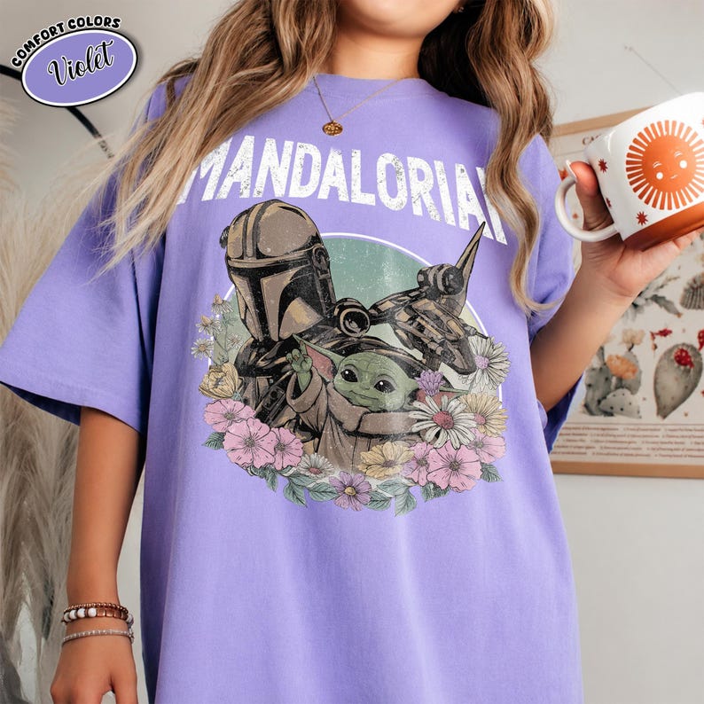 K&ouml;nnte beinhalten: Ein violettes T-Shirt mit einer Grafik des Mandalorianers und Grogu, umgeben von bunten Blumen. Der Text "MANDALORIAN" ist in Wei&szlig; &uuml;ber den Figuren gedruckt. Ein kleines kreisf&ouml;rmiges Etikett oben links zeigt die Aufschrift "COMFORT COLORS Violet".