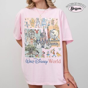 May include: A light pink t-shirt featuring a multi-colored graphic print of various theme park landmarks and cartoon characters. The text "WALT DISNEY WORLD" is printed in blue and orange below the illustration. A "COMFORT COLOR Blossom" logo is visible.