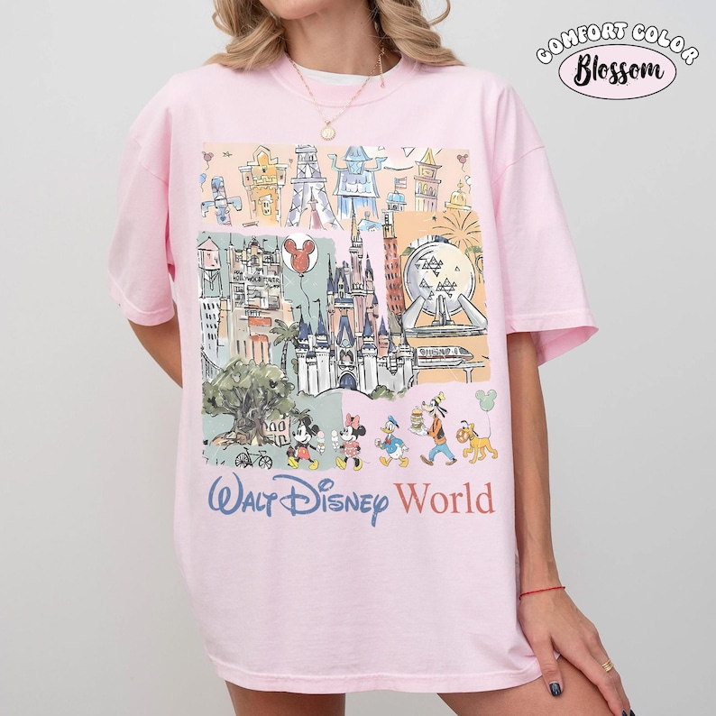 May include: A light pink t-shirt featuring a multi-colored graphic print of various theme park landmarks and cartoon characters. The text "WALT DISNEY WORLD" is printed in blue and orange below the illustration. A "COMFORT COLOR Blossom" logo is visible.