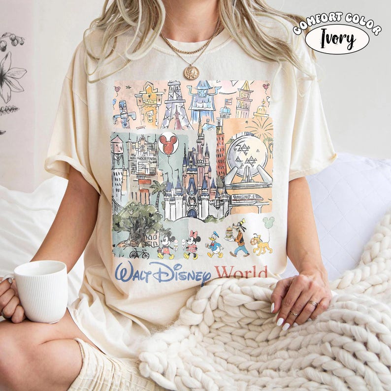 May include: An ivory t-shirt featuring a colorful graphic print of Walt Disney World landmarks and characters including Mickey Mouse, Minnie Mouse, Donald Duck, Goofy, and Pluto. The text reads Walt Disney World and Comfort Color Ivory.