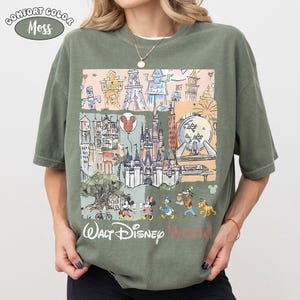 May include: A moss green short-sleeve t-shirt featuring a graphic print of various theme park landmarks, including a castle, a geodesic sphere, and cartoon characters. The text "Walt Disney World" is printed in white and red at the bottom.