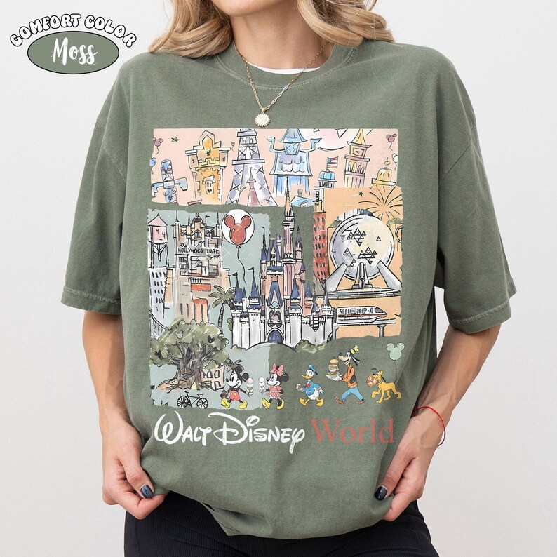 May include: A moss green short-sleeve t-shirt featuring a graphic print of various theme park landmarks, including a castle, a geodesic sphere, and cartoon characters. The text "Walt Disney World" is printed in white and red at the bottom.