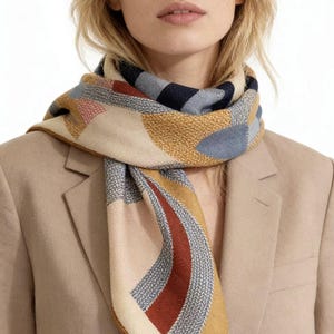 Puede incluir: Bufanda beige con un patrón geométrico en tonos azules, amarillos, rojos y rosas. La bufanda está drapeada alrededor del cuello y los hombros, combinada con una blazer color beige. El diseño de la bufanda presenta una mezcla de formas y texturas.