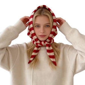 Könnte beinhalten: Ein rot-weiß gestreifter Schal, der um Kopf und Hals gebunden ist. Der Schal ist aus einem weichen Material gefertigt und wird über einem cremefarbenen Pullover getragen. Die Streifen des Schals sind gleichmäßig angeordnet und bilden ein auffälliges Muster.